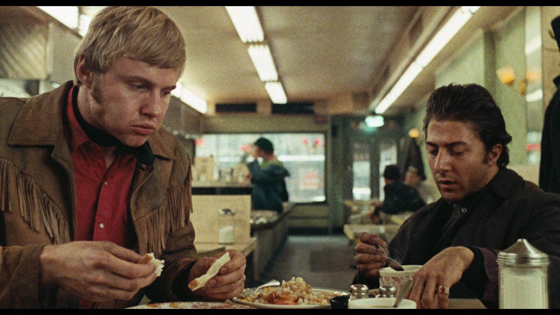Joe and Rico sit in a classic American-style diner. Joe's mouth is full as he pulls apart a piece of toast. He is wearing a red button-up, his signature black neckerchief, and his brown, frilled jacket. Rico is wearing a black shirt with a similarly colored overjacket. He is eating something from a bowl, spoon in hand.
