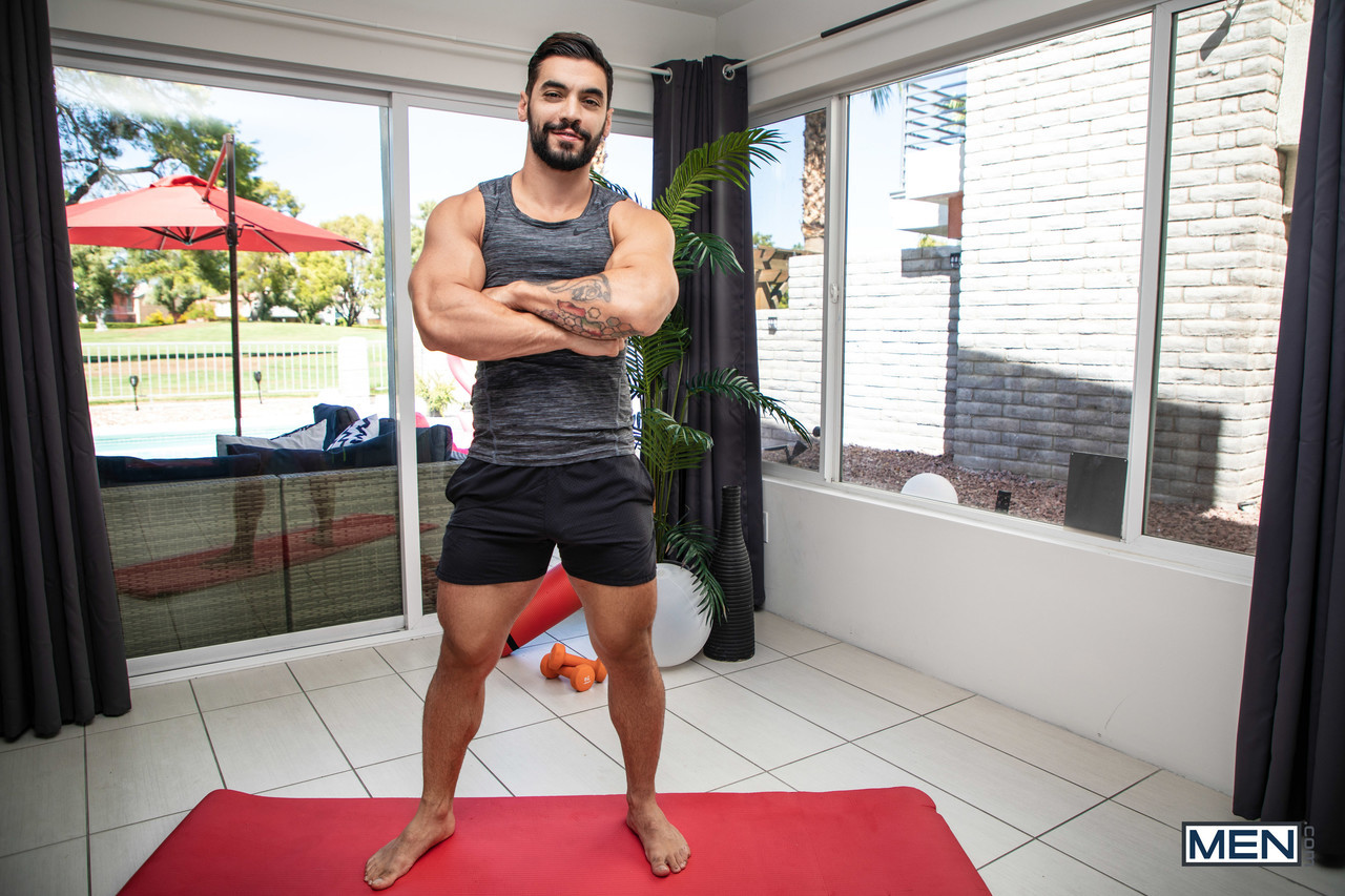 Hunky man with black hair stands on exercise mat with arms crossed wearing gym clothes
