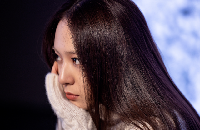 A photo of an asian woman. She has long, straight, brown hair. She is looking sharply off camera, towards the left, with her chin leaning against her right hand. She has on a white knit sweater, and light makeup. She is standing in front of a cloudy blue LCD screen.