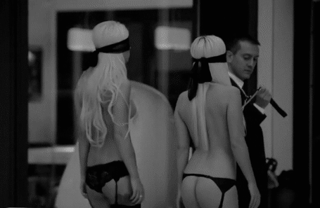 Grayscale image of two blonde women in lingerie on leashes led blindfolded by man in suit