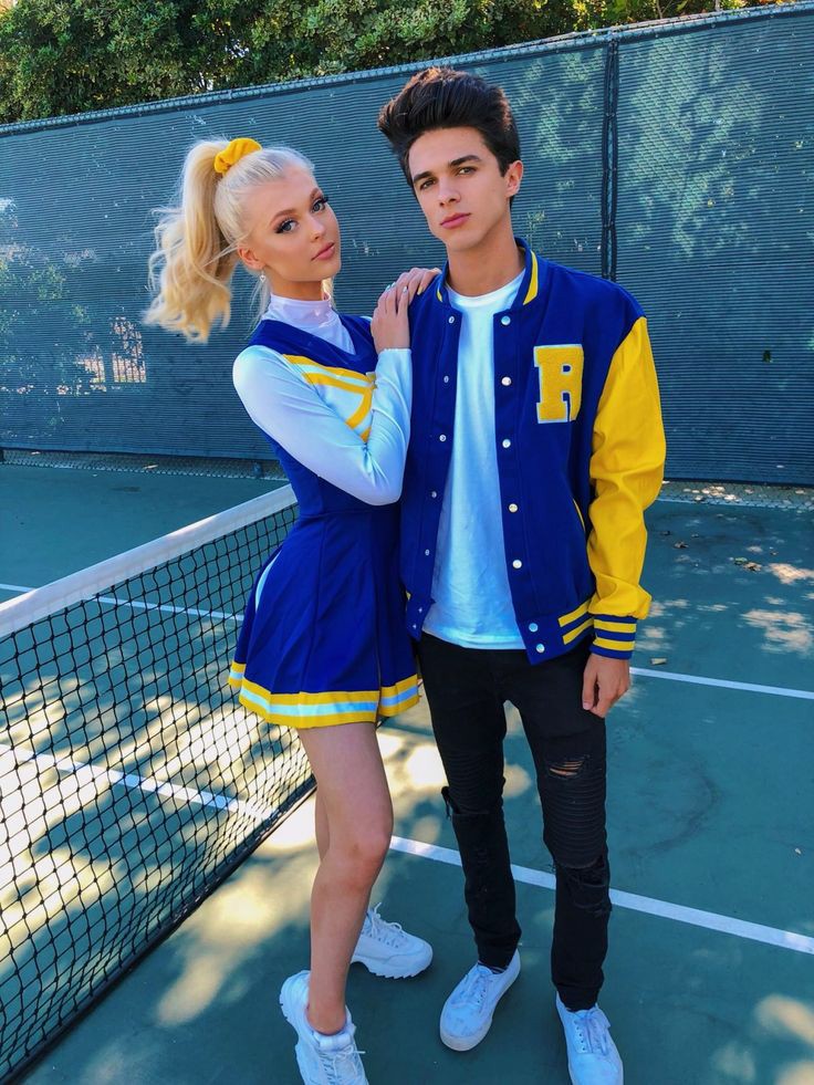 Blonde cheerleader in blue, yellow and white uniform leans on shoulder of dark-haired jock in matching jacket on tennis court