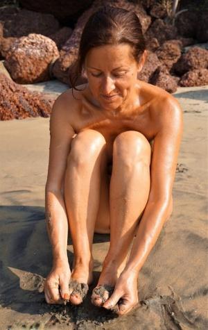 Naked older woman Diana Ananta covers her feet in beach sand at low tide