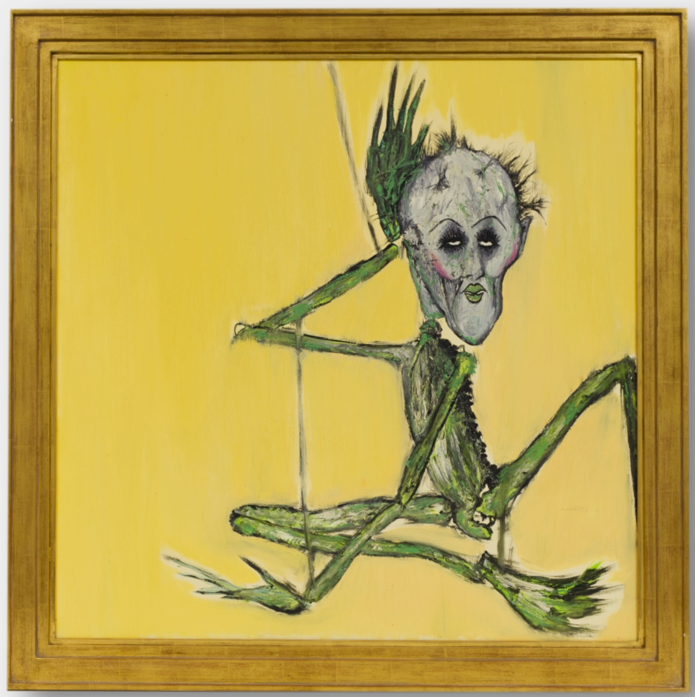 On a plain yellow background there sits a green marionette with patchy hair on its mostly bald head and thick makeup on its male face. One hand is behind its head and the other on the ground, as if posed for a very camp portrait.