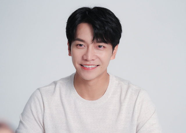 A photo of an asian man. He is sitting in front of a white wall, wearing a white long-sleeved u-neck shirt. His black hair ends at his eyebrows, and his forehead is mostly obscured by his bangs. He is smiling at the camera, and facing slightly towards the left.