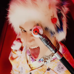 Icon of Griffith. He is wearing a white fur hat with a blue steak, a tie-dye shirt, and has a cane with a plastic eyeball at the top. He is posing with the cane in front of his right eye, and is sticking his tongue out to the side with a fierce and playful expression.