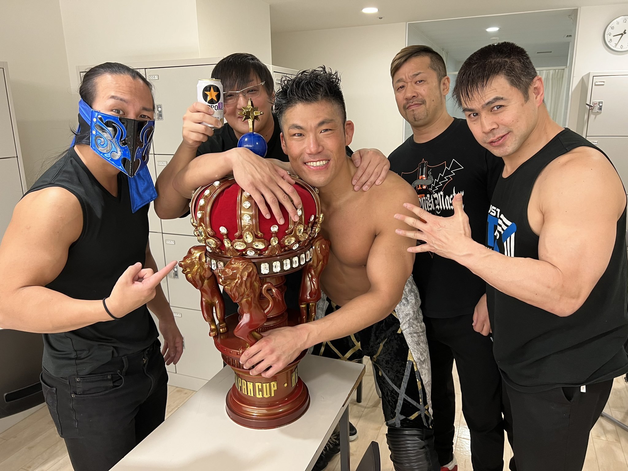 an Image of Sanada smiling with the New Japan Cup Trophy, still in his wrestling gear. Members of Just 5 Guys surround him. everyone is smiling