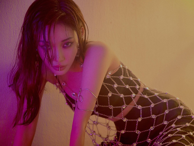A photo of a feminine asian person. Xie is lit by purple lighting and in front of a yellow-brown wall. Xer knees are folded on the ground, and xie's leaning up by xer forearms. Xer hair is wavy and brown, going down past xer bust. Xie is wearing a black two-piece bodysuit, that shows a hint of midriff. Xie has a fake lip-ring on, and a silver arm bangle. Xie also has a dress made of a silver chain, with white jewels connecting the chains. Xie is staring directly into the camera.