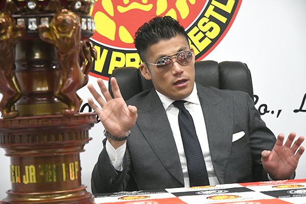 image of Sanada at the Sakura Genesis Press Conference. He is dressed sharply in a black suit with sunglasses. The NJ Cup trophy sits slightly out of focus in the foreground