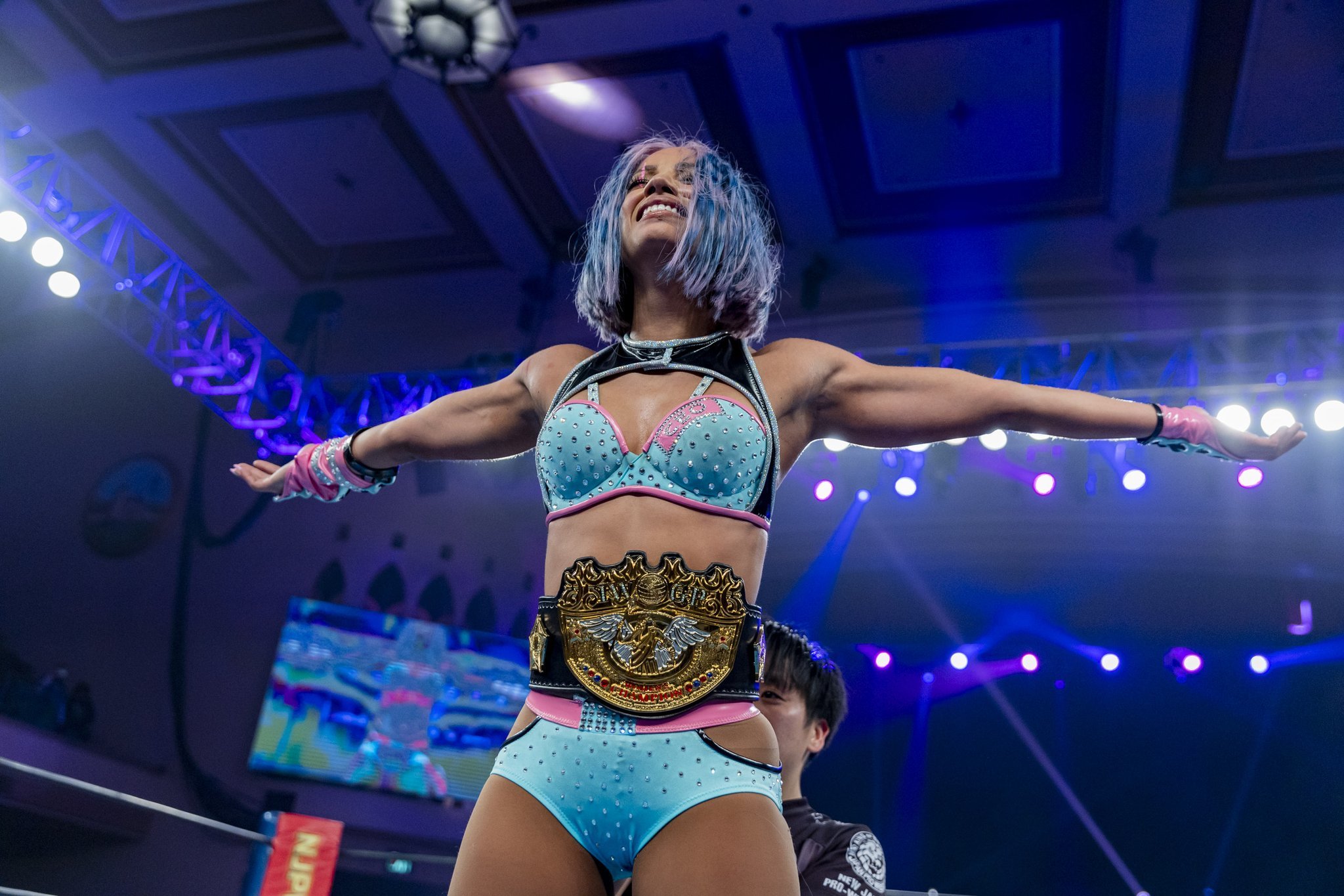 an image of Mercedes Mone after her match against Kairi, standing proudly with her arms outstretched as the IWGP Womens Championship Belt is secured around her waist by a referee.