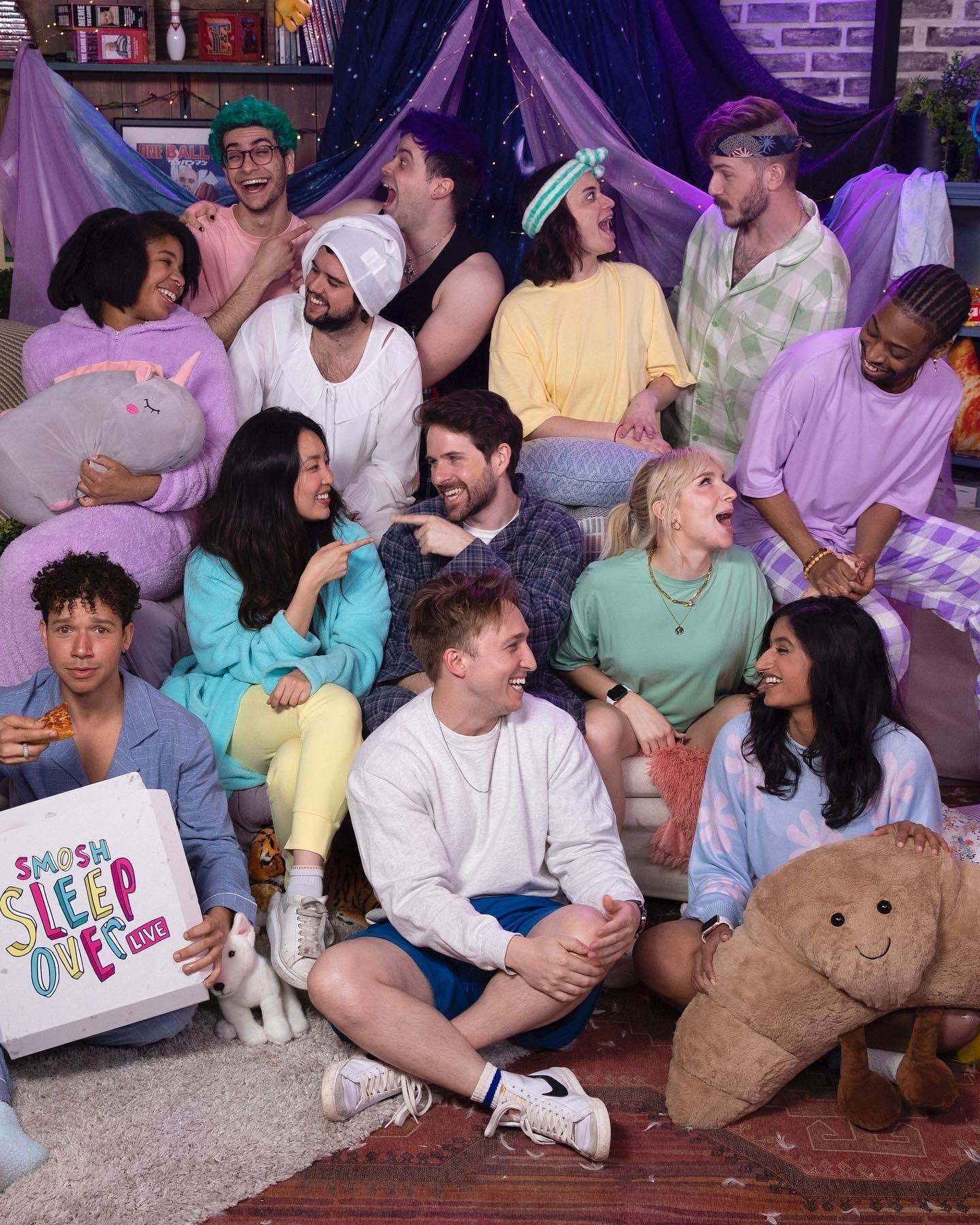 an image of the main smosh cast. they are all wearing pajamas and interacting with each other in a silly fashion. in the bottom left, one cast member is holding a pizza box that reads 'Smosh Sleep Over Live' on the back.