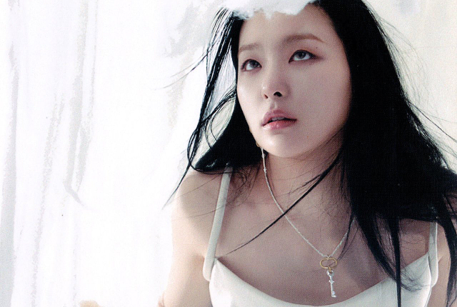 Photo of an asian woman, standing against a white feathered background. She is looking with an innocent facial expression toward the sky. She has long black hair, and a neutral, if quizzical expression on her face. She is wearing a white dress, and a necklace with a large key charm.