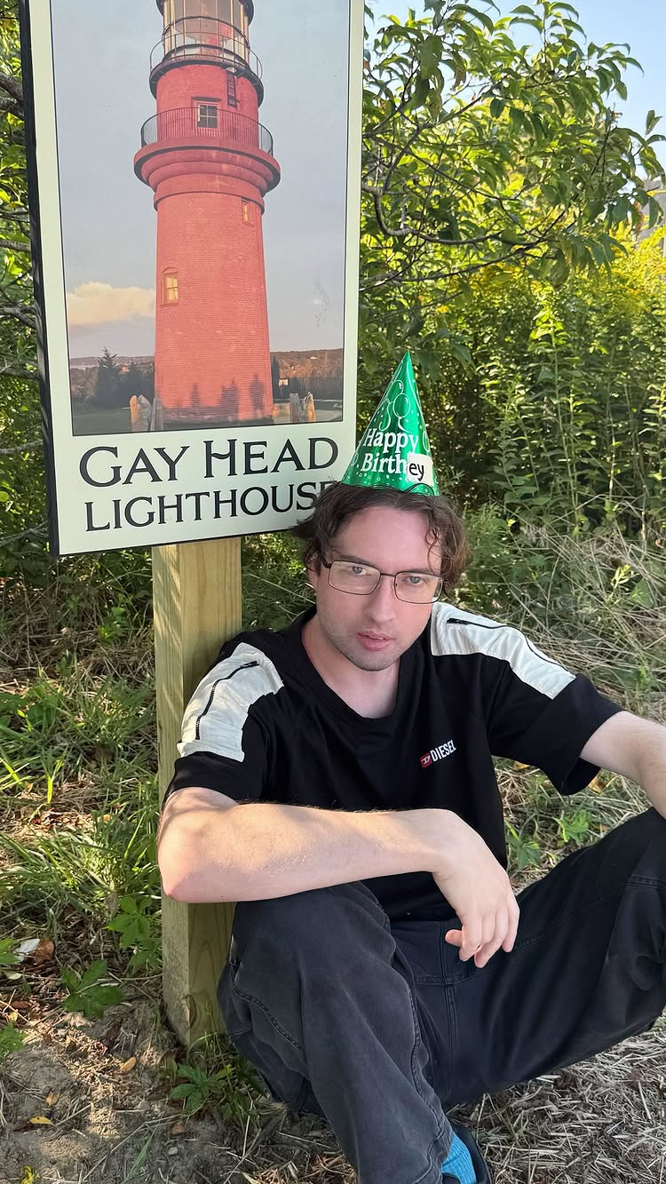 Andrew Hussie sitting in front of a sign for the Gay Head Lighthouse while wearing a birthday hat hastly made to say "birthey" instead