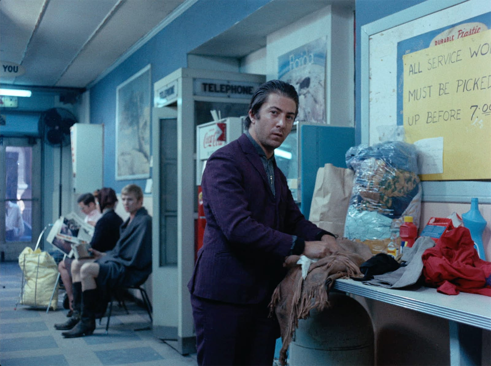 Rico is dressed in a rich purple suit. He leans over a counter in a laundromat, scrubbing at Joe's frilled cowboy jacket. He seems distracted, looking over his shoulder. Joe sits in a in the background, covered by a blanket. A few others sit beside him, holding their laundry.