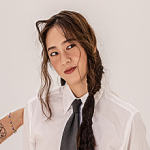 An icon of Pruu. She is smiling with her head tilted, and someone is reaching upwards to fix her long brown hair. She is posing against a white background, and wearing natural makeup with red lipstick.