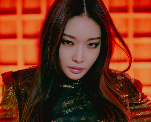 A photo of a feminine non-binary asian person. Xie has long brown hair, and is gazing straight into the camera with a confident, daring expression. Xie is wearing a dark green and black embellished top, and standing in front of a red grid background.