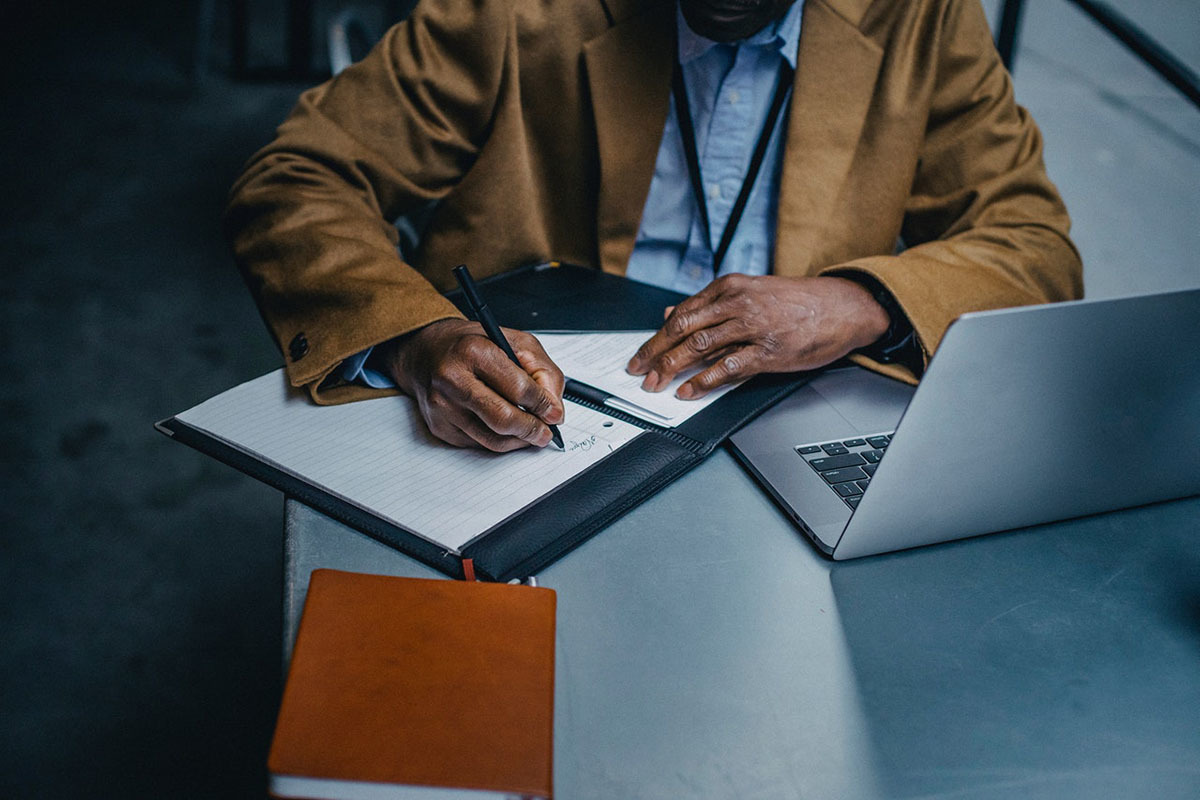 Black male executive writing on paper sheet near laptop