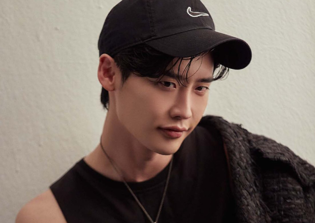 A photo of an asian man. He is standing against a white wall, and wearing a black baseball cap with the Nike logo on it stitched in white. he is wearing a black tank top, with a black knitted jacket hanging off his right shoulder, and a gold necklace. His black bangs are slightly peaking out from the brim of his cap, framing his face. He is smirking and looking towards the right. 