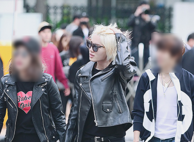 A photo of an asian person of questionable gender. They have blond hair that goes down to their chain, and are wearing a classic leather jacket and tortoiseshell sunglasses. They're smoothing their hair back with their left hand. They have a black shirt underneath and are wearing black pants and a black belt with a silver buckle. They're walking with two other people.
