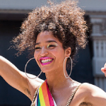 Icon of Dianna. She is smiling and lifting her arms up in a superwoman pose (right arm up and upward, left arm pulled back and flexing). She has on bright makeup, and is wearing a rainbow sash. Her brown, curly, hair is tied up in an updo.