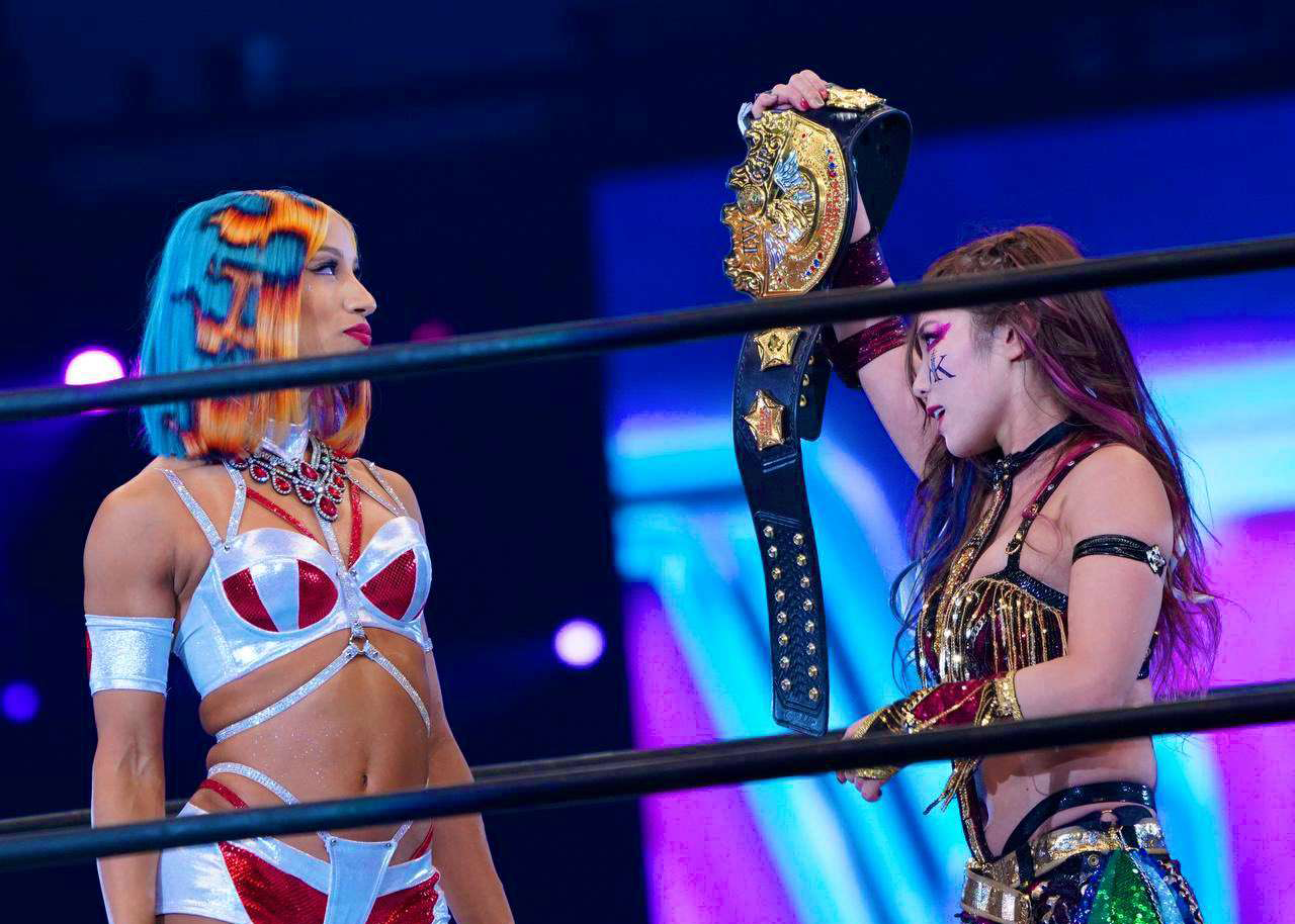 an image of Kairi, expression serious, holding up the IWGP Women's Championship. She staring down Mercedes Mone, who stands across from Kairi. Mone is staring at the championship with a determined, happy expression.