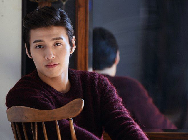 Photo of an asian man with black hair. He is wearing a red sweater, and looking into the camera with a warm expression. He is sitting down in a wooden chair in front of a mirror. He seems about as stiff as the chair.