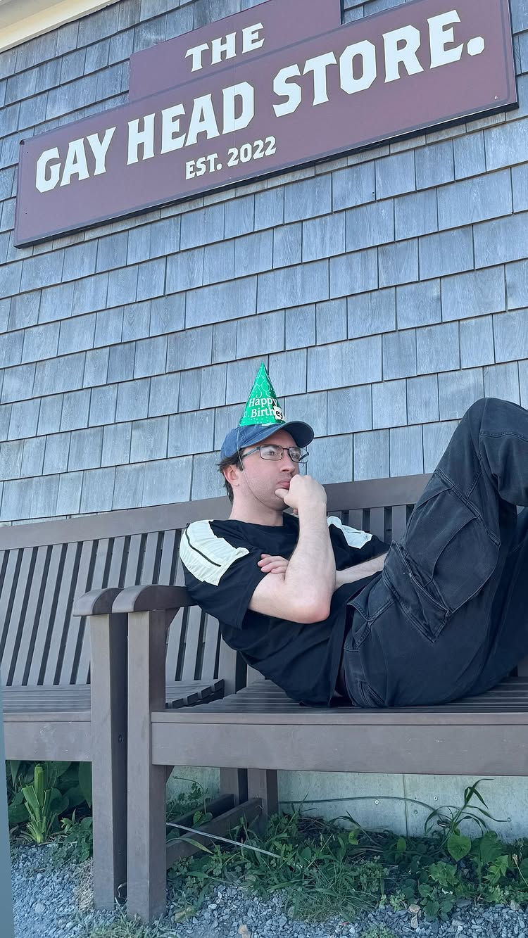 Andrew Hussie looking pensive while chillin on a bench below a sign reading The Gay Head Store est. 2022. The birthey hat is now on top of a different hat.