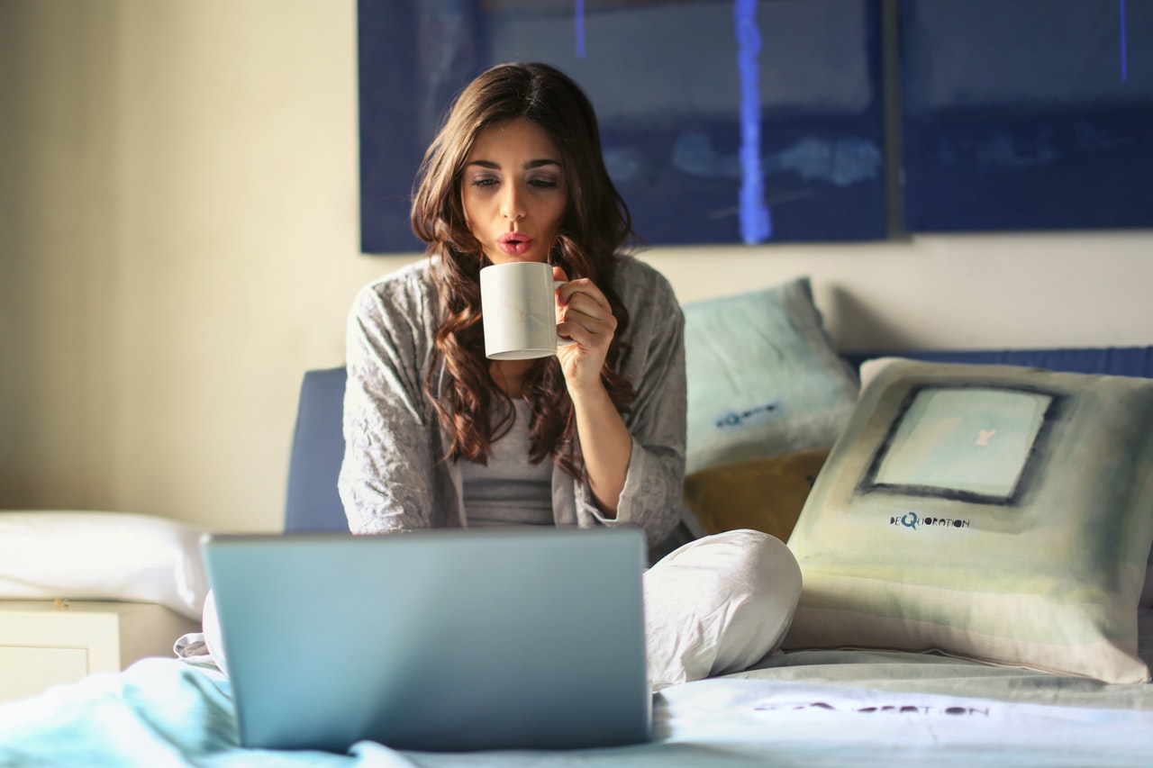Brunette woman sits on bed in pyjamas using laptop and blowing hot drink