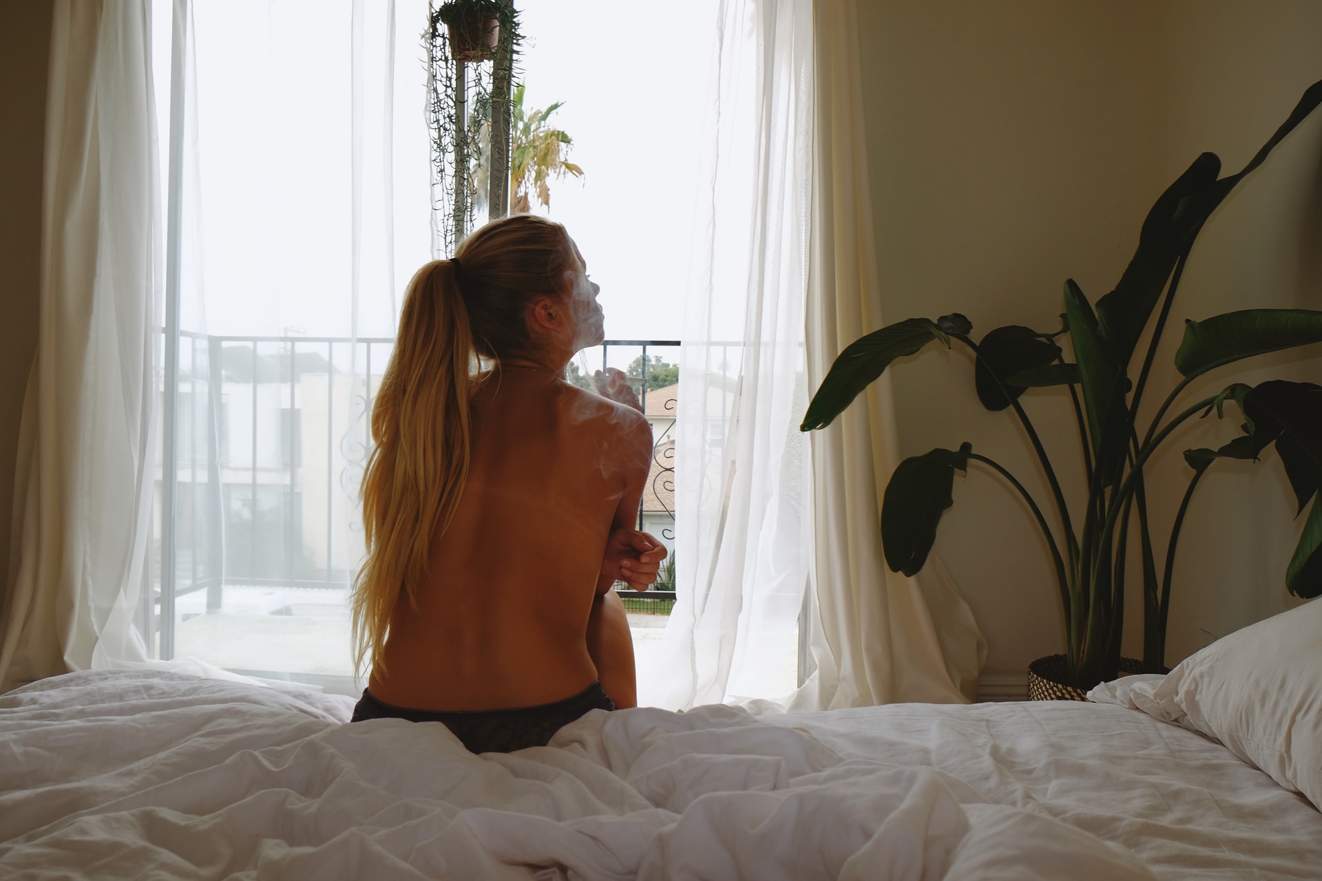 Blonde semi-naked woman sits on bed smoking in front of balcony