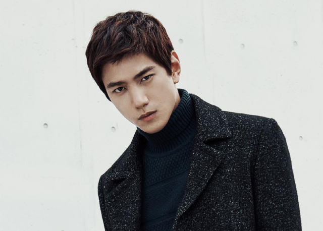 A photo of an asian man with tousled brown hair. He is staring intensely into the camera, and standing against a white background with small grey circles. He is wearing a black coat with white specks, and a black turtleneck underneath. He doesn't seem to be very interested in you.