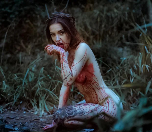 Naked tattooed woman with blood smeared over chest, arms, legs and face kneels on forest floor and licks back of hand