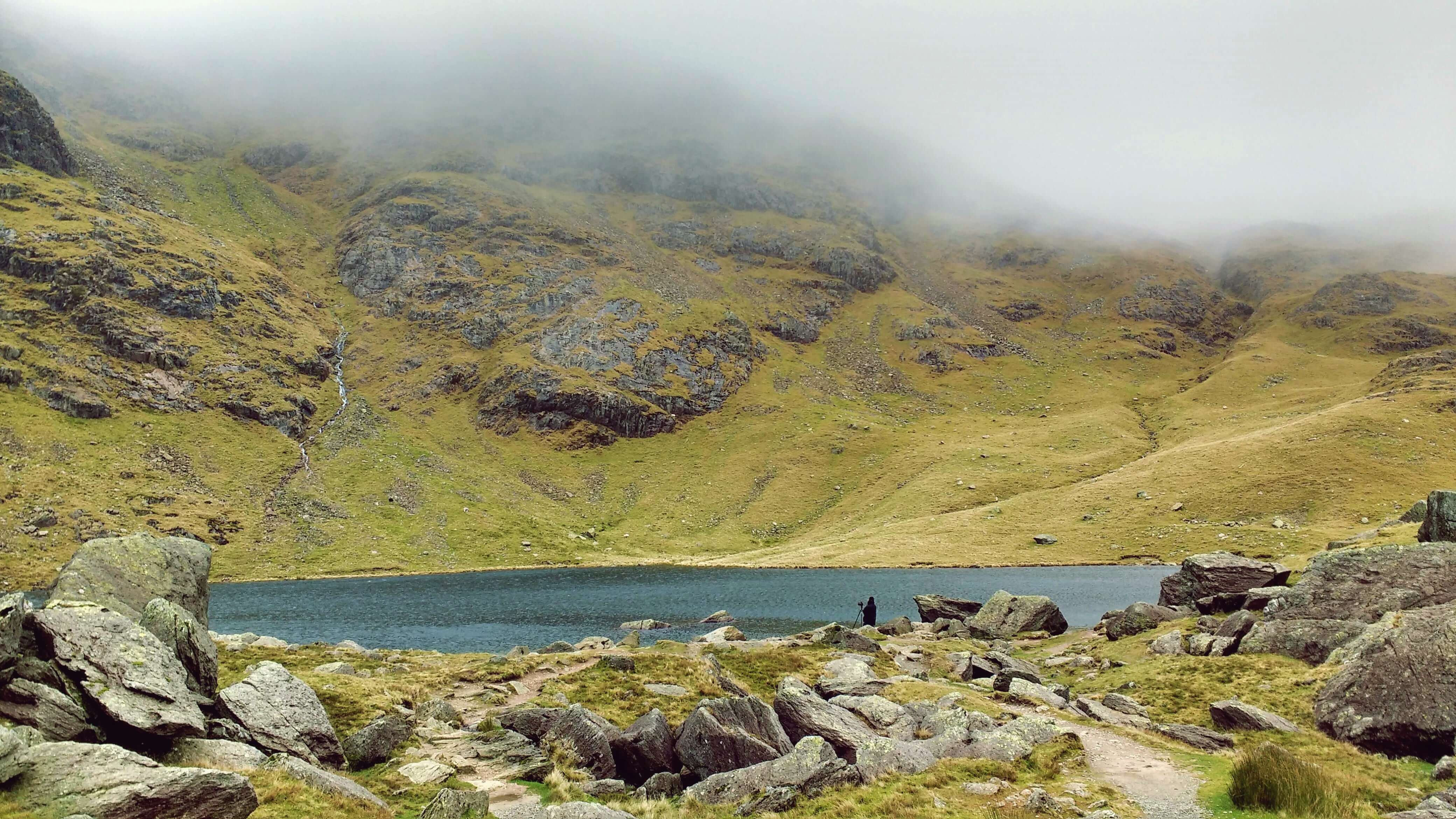 A figure standing by a lake surrounded by rocky fells