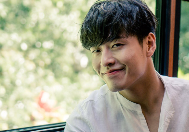 A photo of an asian man. He is sitting down in front of a window, and outside the window there is a lot of greenery. He is looking at the camera at an angle, and smiling a closed-lipped smile. His hair is short, black, and curly. He is wearing a white shirt with the first button unbuttoned. 