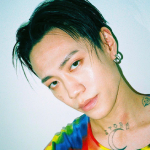 An icon of Jason. He's wearing a rainbow u-neck shirt and standing against a light-blue wall. One of his tattoo's is showing, and his hair is parted with his bangs framing his face.