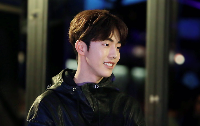 A photo of an asian man. He is looking to the right with a smile on his face. He has brown hair that's split in the middle, with bangs framing his eyebrows. He is wearing a black windbreaker, and standing in front of a dark window, black with hints of blue. 