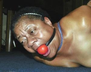 Overweight black woman Trixie struggles against a ball gag while hogtied