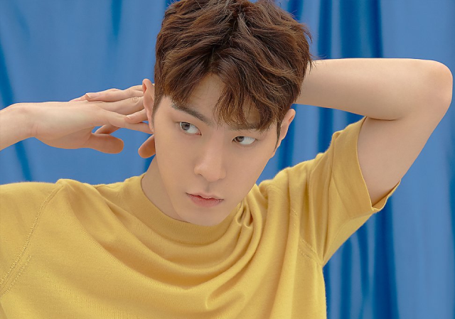 A photo of an asian man. He is standing in front of a blue curtain, and wearing a yellow shirt. His brown hair is short and wavy. He has his hands interlocked behind his head, and is looking off towards the side with a stern, but questioning, expression.