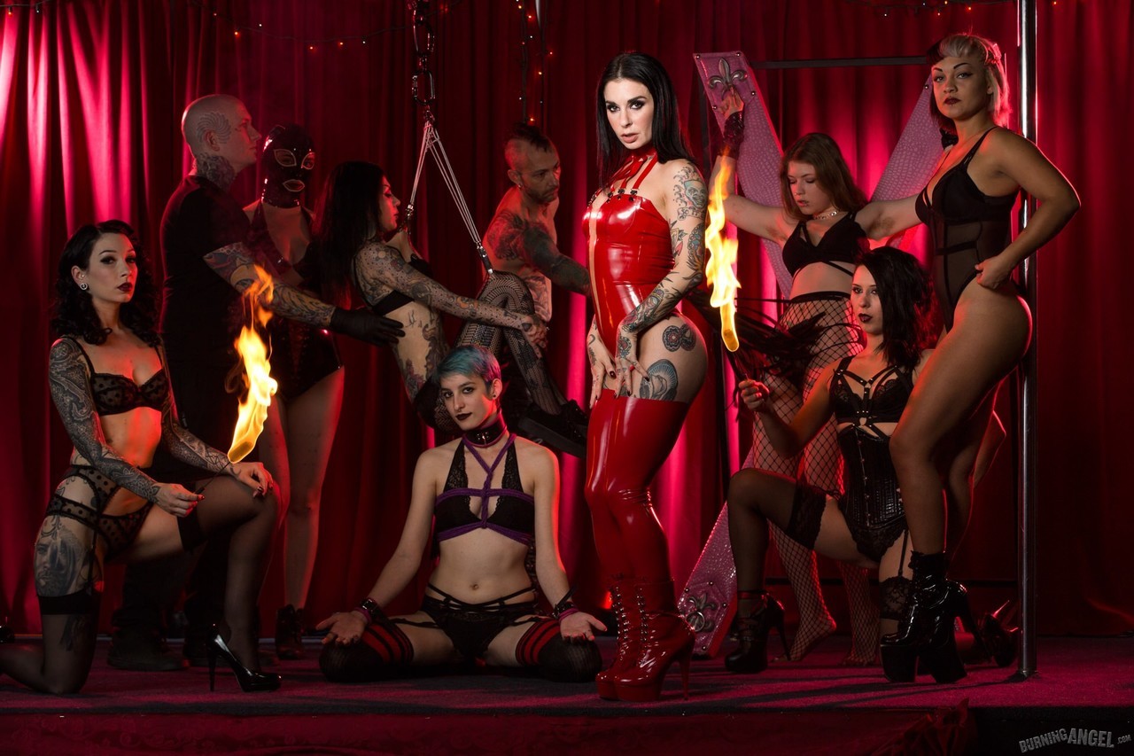 Black-haired tattooed woman in red latex outfit stands amongst group of alt men and women in erotic positions in front of red curtain background