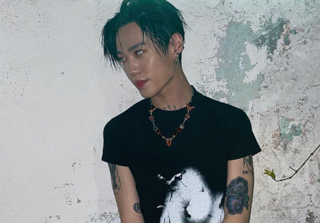 A photo of an asian man. He is standing against a white wall with several green splotches. His hair is black, slicked, and messy, going down past his ears at some points. He is looking off to the left. He has many tattoo's- one is peaking out of the top of his shirt, the others on his arms. He is wearing a black graphic tee with a necklace over it. 